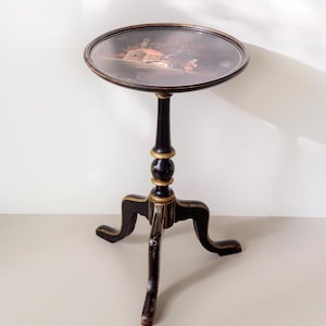 Op de afbeelding: Een zwarte bijzettafel met een rond blad voorzien van een geschilderd landschap. De tafel heeft een centrale kolom met goudkleurige accenten en staat op drie gebogen poten die zijn versierd met fijne goudkleurige lijnen en details.