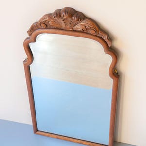 May include: A wall mirror with a medium-brown wooden frame featuring a carved shell motif at the top centre. The mirror has a rectangular shape with curved upper corners and a smooth, reflective glass surface framed by detailed woodwork.