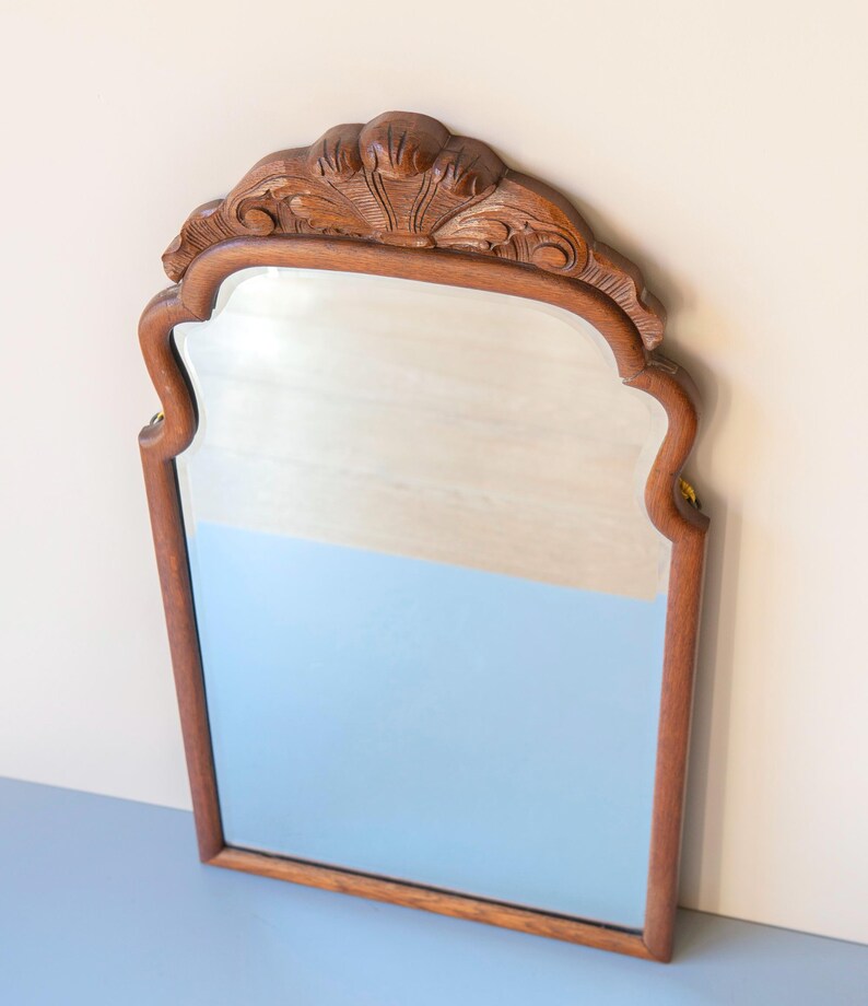 May include: A wall mirror with a medium-brown wooden frame featuring a carved shell motif at the top centre. The mirror has a rectangular shape with curved upper corners and a smooth, reflective glass surface framed by detailed woodwork.