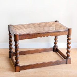 Puede incluir: Un taburete de madera vintage con asiento de mimbre tejido. El marco marrón oscuro presenta patas talladas en espiral y un faldón festoneado. El taburete es una pieza de mobiliario clásica.