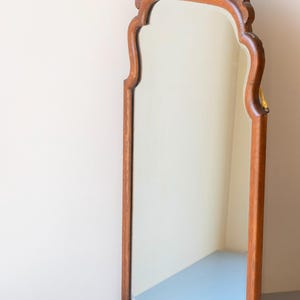 May include: A tall, vertical wall mirror featuring a medium-brown wooden frame with a smooth, polished finish. The top of the frame is arched and decorated with intricate, carved floral and shell-like patterns, adding texture to the wood.