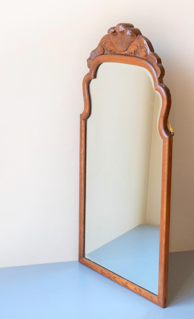 May include: A tall, vertical wall mirror featuring a medium-brown wooden frame with a smooth, polished finish. The top of the frame is arched and decorated with intricate, carved floral and shell-like patterns, adding texture to the wood.