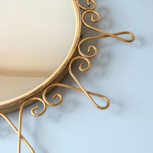 Puede incluir: Espejo redondo dorado con un marco ornamentado en forma de sol. El marco presenta adornos met&aacute;licos en espiral. El espejo est&aacute; sobre un fondo azul claro, creando una est&eacute;tica vintage.