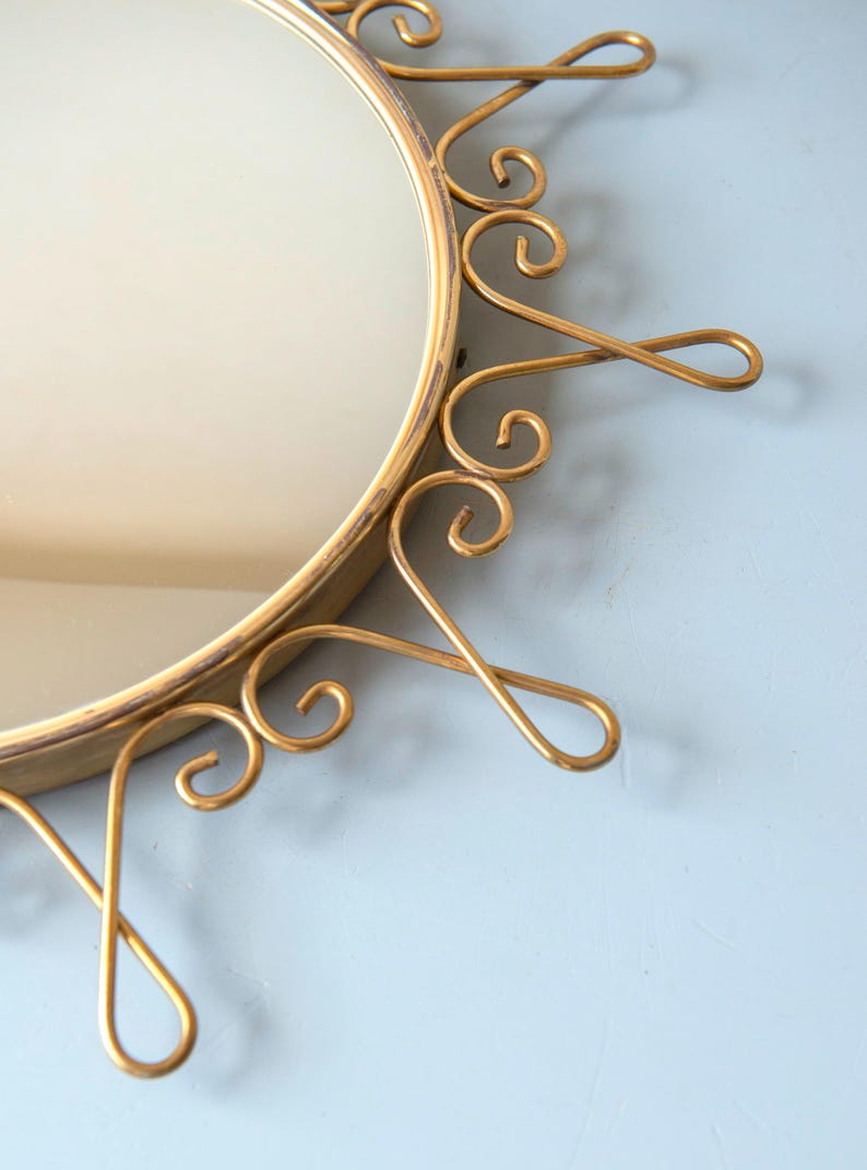Puede incluir: Espejo redondo dorado con un marco ornamentado en forma de sol. El marco presenta adornos met&aacute;licos en espiral. El espejo est&aacute; sobre un fondo azul claro, creando una est&eacute;tica vintage.