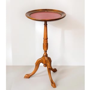 Peut inclure: Une table piédestal en bois dotée d'un plateau circulaire avec un rebord surélevé et une incrustation en cuir rouge foncé. La table repose sur une colonne centrale fine et tournée, soutenue par trois pieds incurvés aux extrémités arrondies.