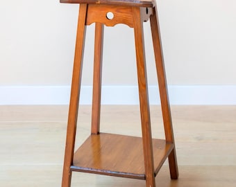 May include: A tall, wooden plant stand with a rectangular top and a lower shelf. The stand has four angled legs and a decorative cutout near the top. The wood has a warm, reddish-brown tone, and the stand is approximately 30 inches tall.