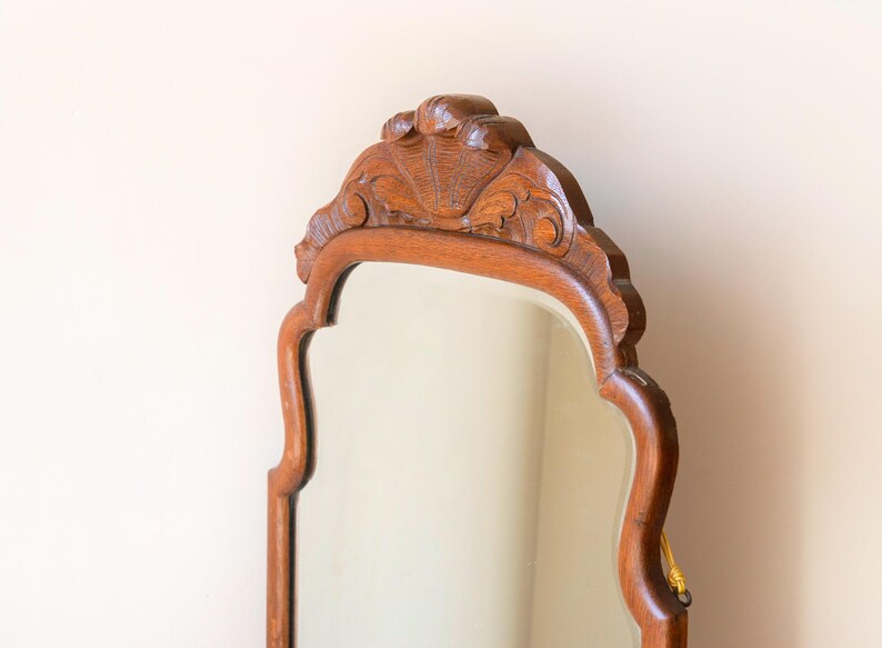 May include: A wall mirror with a medium-brown wooden frame featuring a curved, scalloped silhouette. The top of the frame is adorned with a carved shell motif and decorative scrollwork. A small yellow cord is visible on the right side of the frame.