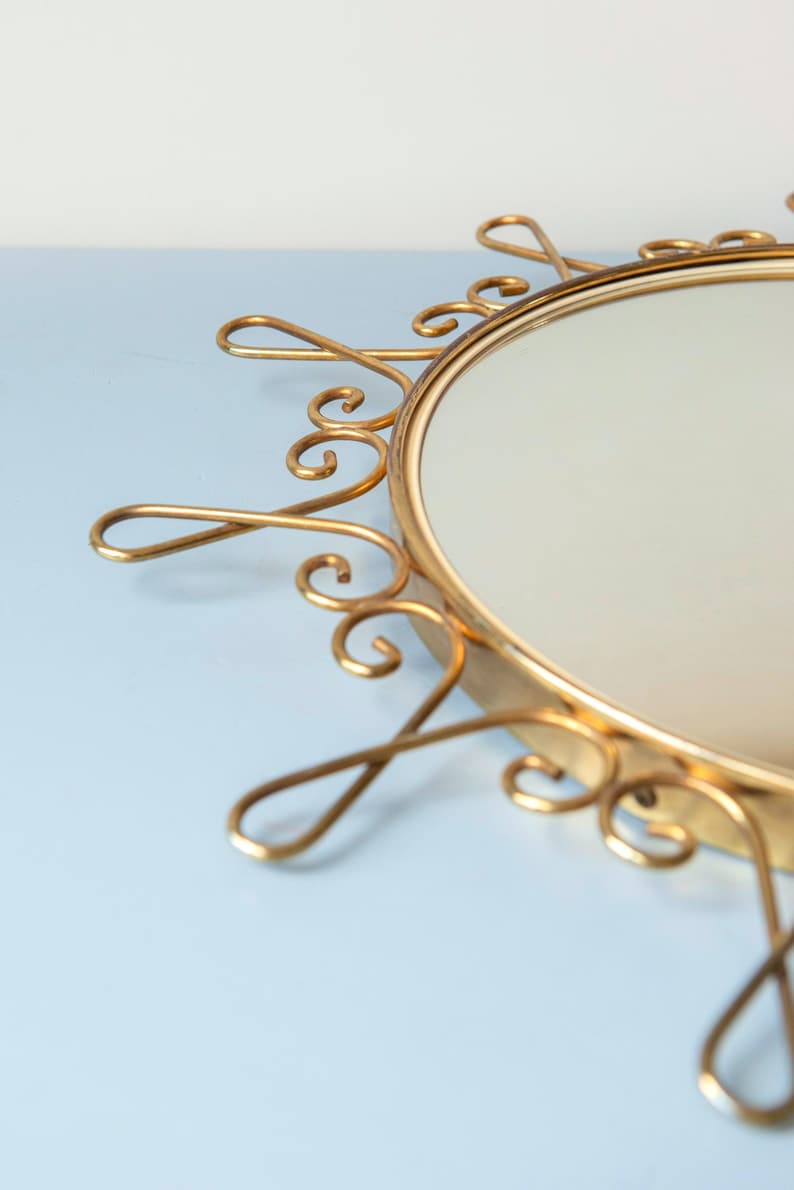 Puede incluir: Espejo redondo dorado con un marco ornamental en forma de sol. El marco presenta delicados adornos met&aacute;licos en espiral alrededor del borde del espejo. El espejo est&aacute; sobre un fondo azul claro.