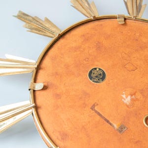 May include: A vintage sunburst mirror with a gold-tone metal frame and radiating rays. The mirror's back is a warm, reddish-brown colour, and it features a small, circular emblem with text. A decorative piece for wall decor.