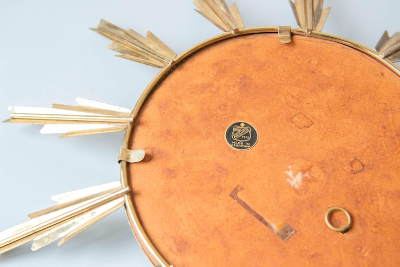 May include: A vintage sunburst mirror with a gold-tone metal frame and radiating rays. The mirror's back is a warm, reddish-brown colour, and it features a small, circular emblem with text. A decorative piece for wall decor.