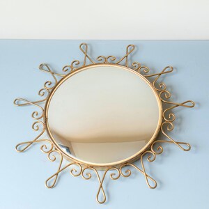 Puede incluir: Espejo redondo dorado con dise&ntilde;o de sol. La superficie reflectante es circular, rodeada por un marco met&aacute;lico adornado con espirales y bucles decorativos. El dise&ntilde;o es elegante y llamativo.