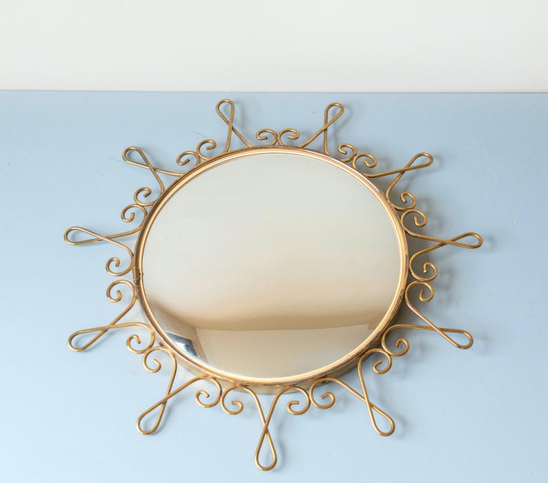 Puede incluir: Espejo redondo dorado con dise&ntilde;o de sol. La superficie reflectante es circular, rodeada por un marco met&aacute;lico adornado con espirales y bucles decorativos. El dise&ntilde;o es elegante y llamativo.