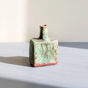Op de afbeelding: Een kleine, vierkante keramische vaas met een getextureerd oppervlak. De vaas is voornamelijk groen met roodbruine accenten en een rode basis. De hals van de vaas is cilindrisch en heeft een kleine opening.