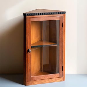 Puede incluir: Un mueble de esquina de madera con puerta de cristal. El mueble está hecho de madera de tonos cálidos y presenta una moldura negra en la parte superior. Tiene un pequeño pomo redondo de madera y dos estantes en el interior. El mueble está diseñado para encajar en una esquina.