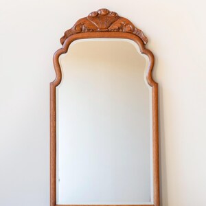 May include: A vertical wall mirror featuring a medium-toned wooden frame with a smooth, rectangular glass surface. The top of the frame is arched with a decorative, carved wooden crest that displays a symmetrical, shell-like floral motif.