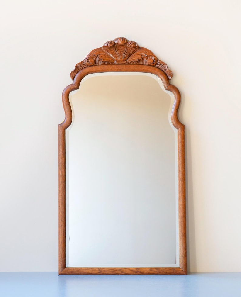 May include: A vertical wall mirror featuring a medium-toned wooden frame with a smooth, rectangular glass surface. The top of the frame is arched with a decorative, carved wooden crest that displays a symmetrical, shell-like floral motif.