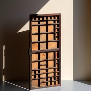 Op de afbeelding: Een rechthoekige houten letterbak met een raster van talloze kleine, open vakjes van verschillende afmetingen. Het frame is donkerbruin, terwijl de interne verdelers en achterpanelen een lichtere, verweerde houten afwerking hebben.