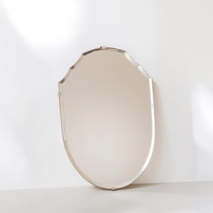 May include: A frameless wall mirror with a shield-like shape featuring a curved top and tapered bottom. The glass has beveled edges that catch the light, and it is held in place by small metal clips positioned at the top, sides, and bottom edges.