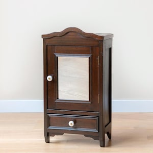 Peut inclure: Une armoire en bois brun foncé avec une seule porte et un tiroir. La porte a une petite poignée blanche et un miroir à l'intérieur. Le tiroir a également une poignée blanche.