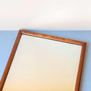 May include: A rectangular wall mirror featuring a warm-toned wooden frame with a visible grain pattern. The mirror glass is clear and reflective, set within the simple, clean-lined wooden border that surrounds the entire perimeter of the piece.