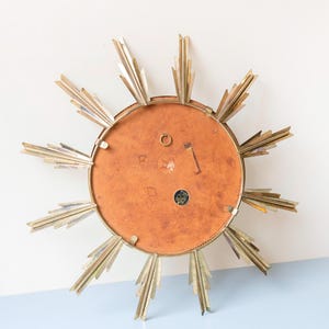 May include: A vintage sunburst mirror with a gold-tone metal frame and radiating rays. The mirror's back is a warm, reddish-brown colour, and it has a small hanging loop. The sunburst design is a classic mid-century modern decor element.