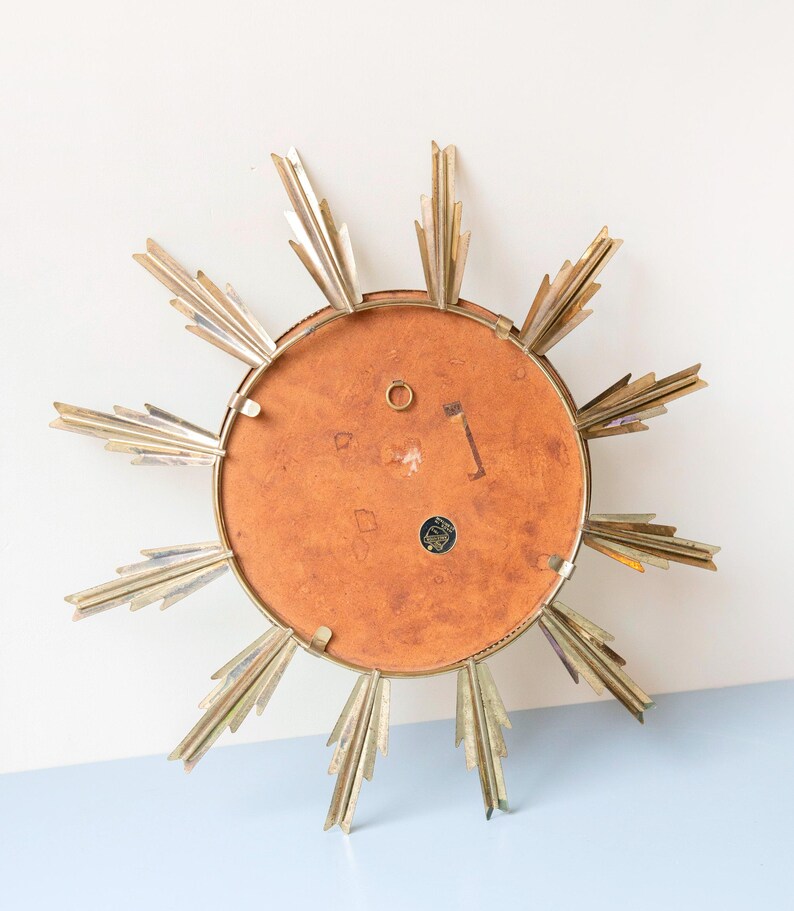 May include: A vintage sunburst mirror with a gold-tone metal frame and radiating rays. The mirror's back is a warm, reddish-brown colour, and it has a small hanging loop. The sunburst design is a classic mid-century modern decor element.