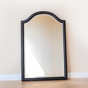 Op de afbeelding: Een hoge, rechthoekige wandspiegel met een glad zwart frame en een opvallende boogvormige bovenkant. Het glazen oppervlak is helder en reflecterend en toont een lichte, neutrale binnenruimte. De spiegel staat rechtop tegen een muur.