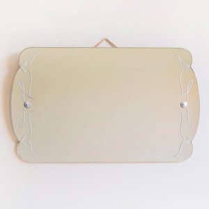 Op de afbeelding: Een rechthoekige spiegel met afgeronde hoeken en decoratieve geëtste lijnen. De spiegel is lichtbeige van kleur en is aan een witte muur bevestigd met een beige touwtje om op te hangen. Twee zilverkleurige klinknagels bevestigen de spiegel.