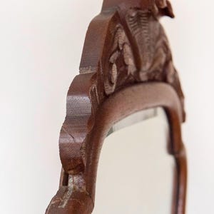 May include: A dark brown wooden mirror frame featuring intricate carved details along the top crest. The frame has a smooth, polished finish and a curved silhouette, showcasing the natural wood grain and the depth of the carved patterns.