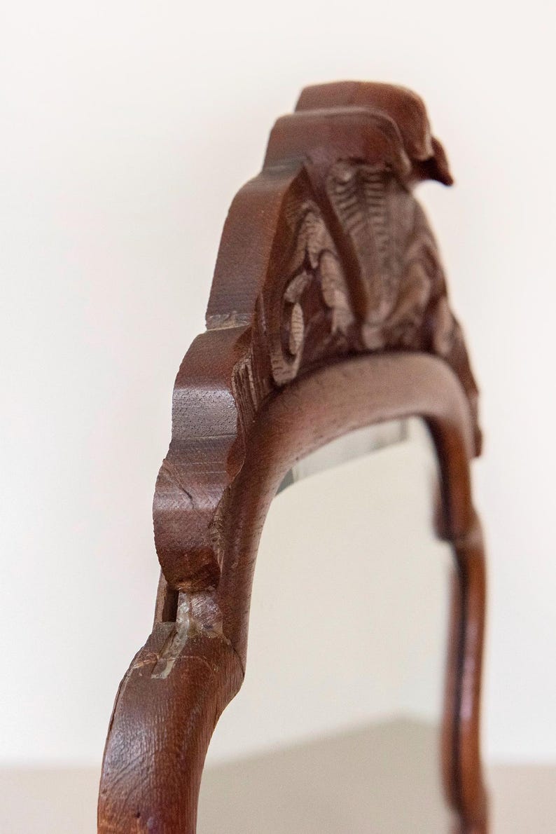 May include: A dark brown wooden mirror frame featuring intricate carved details along the top crest. The frame has a smooth, polished finish and a curved silhouette, showcasing the natural wood grain and the depth of the carved patterns.