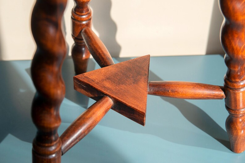 May include: A brown wooden stool base featuring three spiraled legs connected by a central triangular support piece. The legs are cylindrical with a twisted design, and the horizontal supports meet at a flat, solid wood triangle in the center.