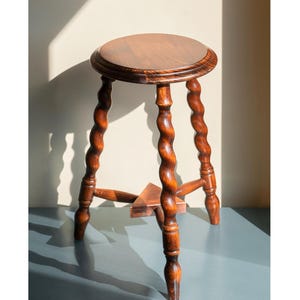 May include: A round wooden stool with a smooth, polished top and three legs featuring a distinct spiral, twisted design. The legs are connected near the base by a small, triangular wooden support piece, all finished in a warm, medium-brown wood tone.