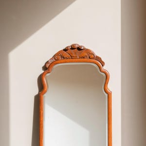 May include: A rectangular wall mirror with a medium-toned wooden frame. The top of the frame features a carved, fan-shaped decorative element. The mirror has a smooth, reflective glass surface and a curved, arched top edge design.