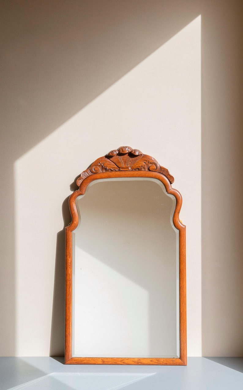 May include: A rectangular wall mirror with a medium-toned wooden frame. The top of the frame features a carved, fan-shaped decorative element. The mirror has a smooth, reflective glass surface and a curved, arched top edge design.