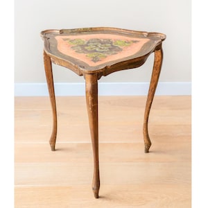 May include: An antique, three-legged side table with a triangular top. The table features a painted floral design in green and brown on a peach-colored background. The table's legs are curved and made of wood.