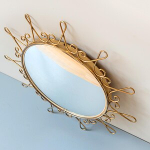 Puede incluir: Espejo ovalado dorado con un marco de metal adornado y arremolinado. El espejo tiene una superficie reflectante y est&aacute; montado en una pared. El marco presenta adornos decorativos, a&ntilde;adiendo una est&eacute;tica vintage. Un art&iacute;culo de decoraci&oacute;n del hogar.