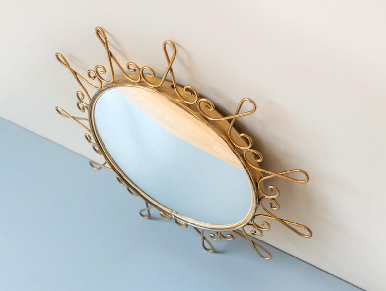 Puede incluir: Espejo ovalado dorado con un marco de metal adornado y arremolinado. El espejo tiene una superficie reflectante y est&aacute; montado en una pared. El marco presenta adornos decorativos, a&ntilde;adiendo una est&eacute;tica vintage. Un art&iacute;culo de decoraci&oacute;n del hogar.