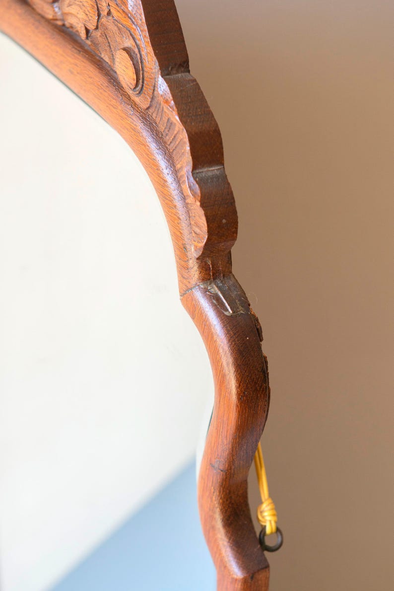 May include: A wooden mirror frame with a medium brown finish and carved decorative details along the top edge. The frame features a curved, scalloped silhouette and includes a yellow cord attached to a metal loop on the side for hanging purposes.