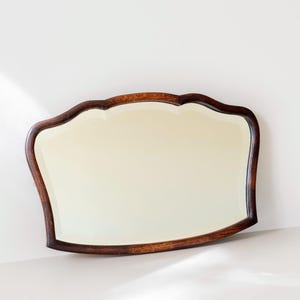 Op de afbeelding: Een wandspiegel met een gepolijst donkerbruin houten frame met een gebogen, geschulpte bovenrand. Het glas is helder en reflecterend, geplaatst in een glad, gecontourneerd frame dat de vorm van de spiegel volgt, wat zorgt voor een breed, horizontaal profiel.