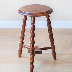May include: A brown wooden stool featuring a circular top and three legs with a twisted, spiral design. The legs are connected near the base by a small, triangular wooden support piece, providing stability to the structure of the furniture.