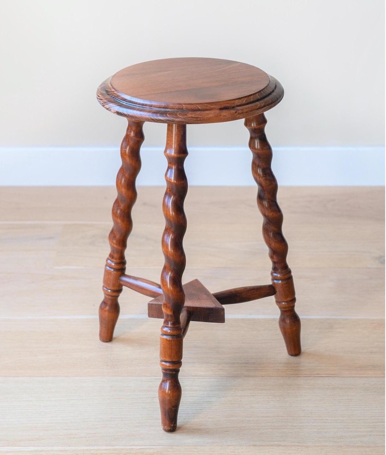 May include: A brown wooden stool featuring a circular top and three legs with a twisted, spiral design. The legs are connected near the base by a small, triangular wooden support piece, providing stability to the structure of the furniture.