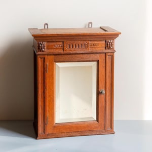 Op de afbeelding: Een houten medicijnkast met een spiegeldeur. De kast is rechthoekig met een decoratieve bovenkant en sierlijke snijwerk. Het hout heeft een warme, roodbruine tint. De kast is ontworpen om aan de muur te worden bevestigd.