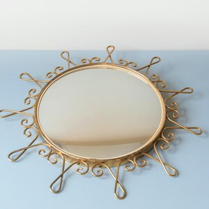Puede incluir: Espejo redondo dorado con un dise&ntilde;o decorativo de rayos de sol. El espejo tiene una superficie reflectante circular rodeada por un marco con acentos arremolinados y en bucle. El marco es de un material met&aacute;lico brillante.