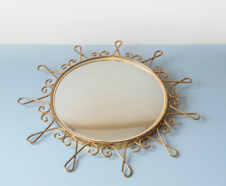 Puede incluir: Espejo redondo dorado con un dise&ntilde;o decorativo de rayos de sol. El espejo tiene una superficie reflectante circular rodeada por un marco con acentos arremolinados y en bucle. El marco es de un material met&aacute;lico brillante.