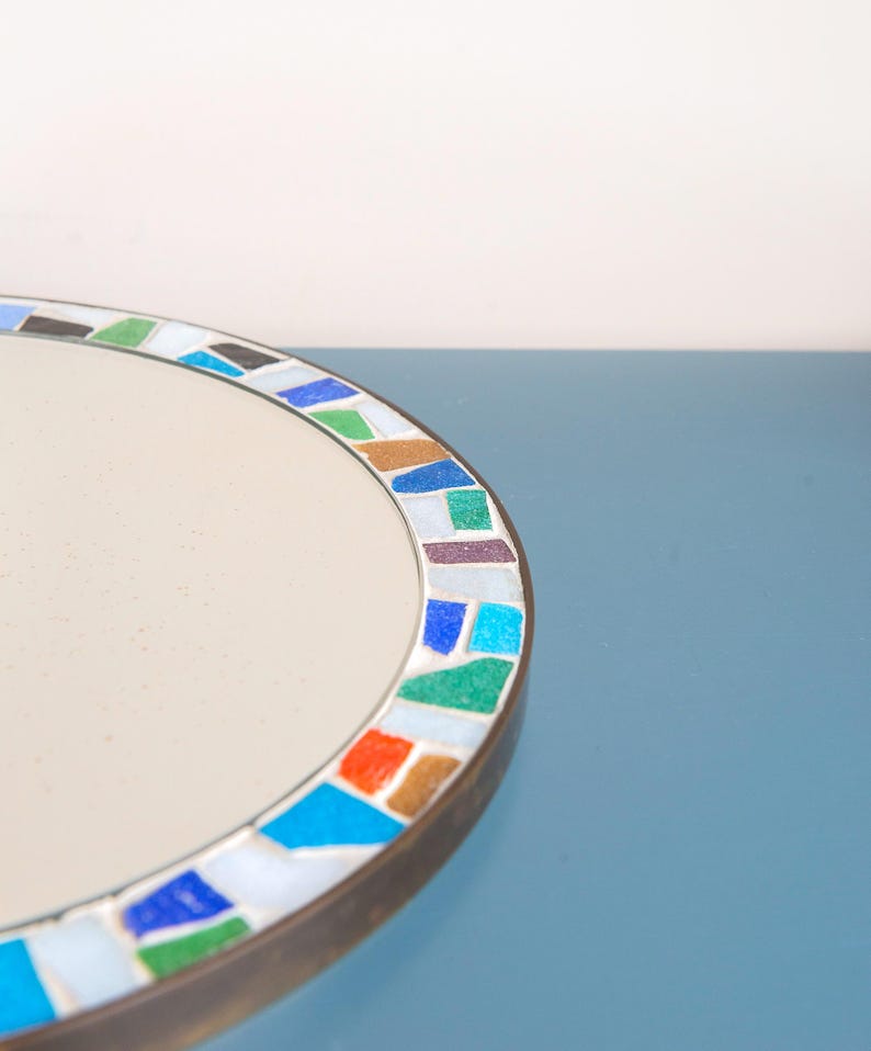 May include: A round mirror with a white surface and a colorful mosaic border. The mosaic border features blue, green, orange, and brown tiles.