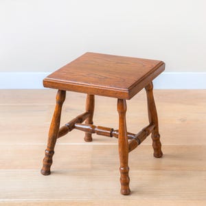 Op de afbeelding: Een kleine, vierkante houten kruk met een rijke, bruine afwerking. De kruk heeft vier gedraaide poten die zijn verbonden door horizontale steunen en is ongeveer 30 cm hoog. De bovenkant is een massief, rechthoekig stuk hout.