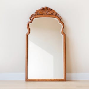 May include: A wall mirror with a medium-toned wooden frame featuring a carved shell motif at the top centre. The mirror has a rectangular shape with rounded upper corners and a smooth, reflective glass surface set within the wooden border.