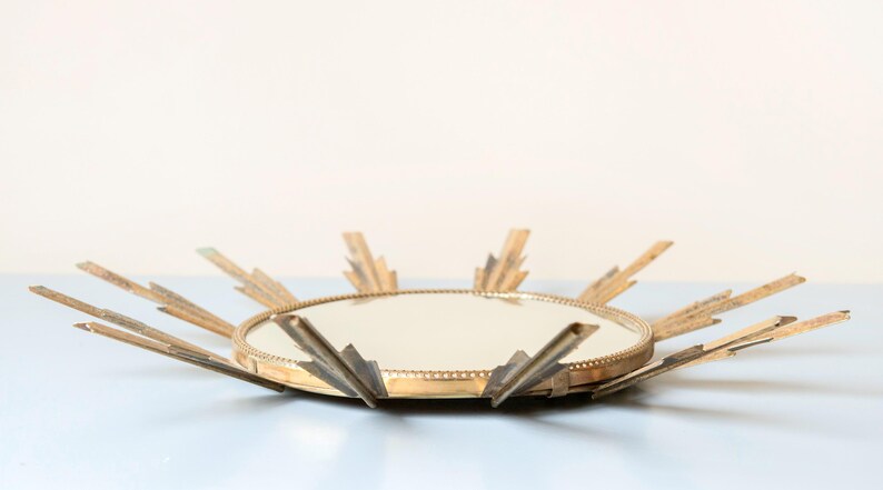 May include: A vintage sunburst mirror with a round mirrored centre and radiating gold-toned metal rays. The mirror has a decorative, mid-century modern design, suitable for wall decor. The overall diameter is approximately 46 cm.