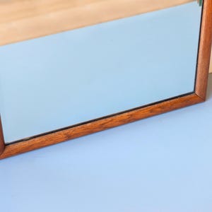 May include: A rectangular wall mirror featuring a medium-toned brown wooden frame with a visible grain pattern. The reflective glass surface is clear and smooth, set within the simple, straight-edged wooden border that surrounds the entire perimeter.