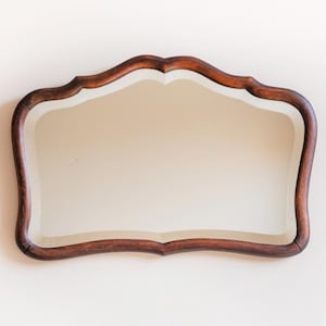 Könnte beinhalten: Ein dekorativer Wandspiegel mit einem dunkelbraunen, verzierten Holzrahmen. Der Spiegel hat eine einzigartige, wellenförmige Form und eine klare, reflektierende Oberfläche. Die satte Farbe und das detaillierte Design des Rahmens verleihen dem Stück Eleganz.